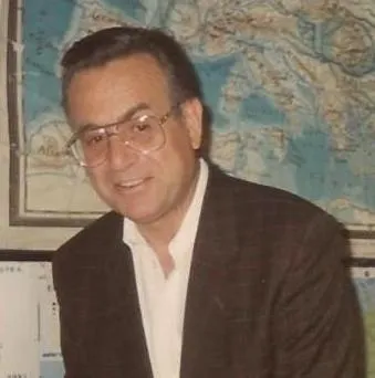 Fotografia d'archivio a colori: Giovanni Ferrari in primo piano, sorridente, in giacca scura e camicia bianca aperta, davanti a una carta geografica appesa alla parete