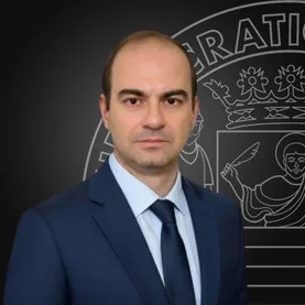Fotografia di Giovannipaolo Ferrari: ritratto frontale a mezzo busto in giacca e cravatta, su fondo scuro con lo stemma dell'ateneo in filigrana