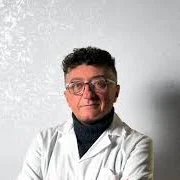 Fotografia di Nikola Tsesmelis: ritratto in piedi a braccia conserte, camice bianco, su fondo chiaro