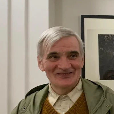 Fotografia di Pascal Duthoit: ritratto in piedi in una sala d'esposizione, giacca verde e maglione color senape, davanti a stampe fotografiche incorniciate